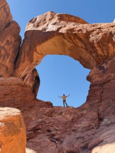 Man with arms raised standing beneath a red rock arch in the Windows section of Arches National Park, Utah